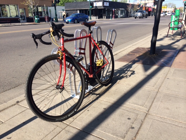 Front, left side view of a red Surly Pacer bike, leaning along a bike rack on a sidewalk with a street behind