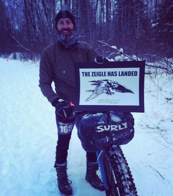 A cyclist standing in snow, next to a fat bike with a Surly front bag, holding a The Zeigle has landed sign