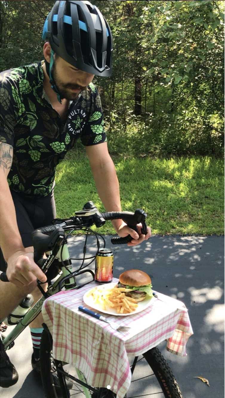 A cyclist standing over a Surly bike on a paved trail with trees, looks down at a plate of food on the bike's gear rack