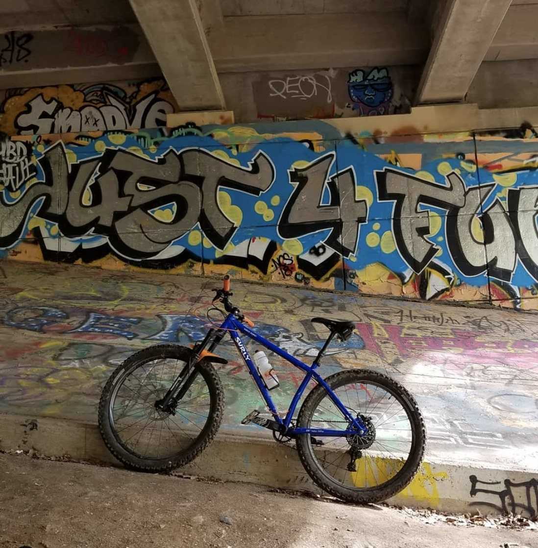 Left side profile view of a blue Surly Karate Monkey bike facing uphill under a graffiti clad concrete bridge