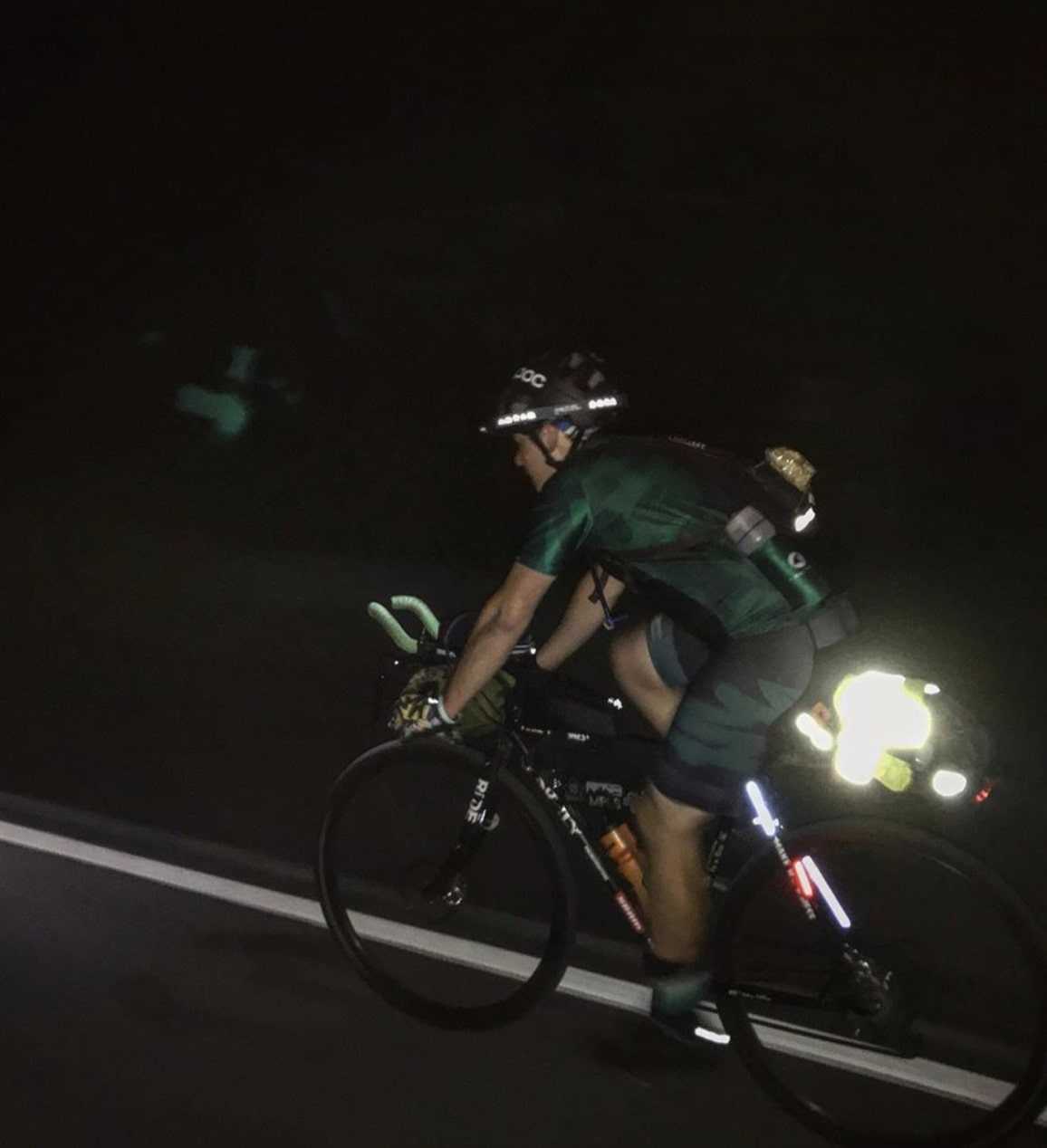 Left side view of a cyclist riding a bike with a reflective rear gear bag pedals in the dark down a paved roadway 