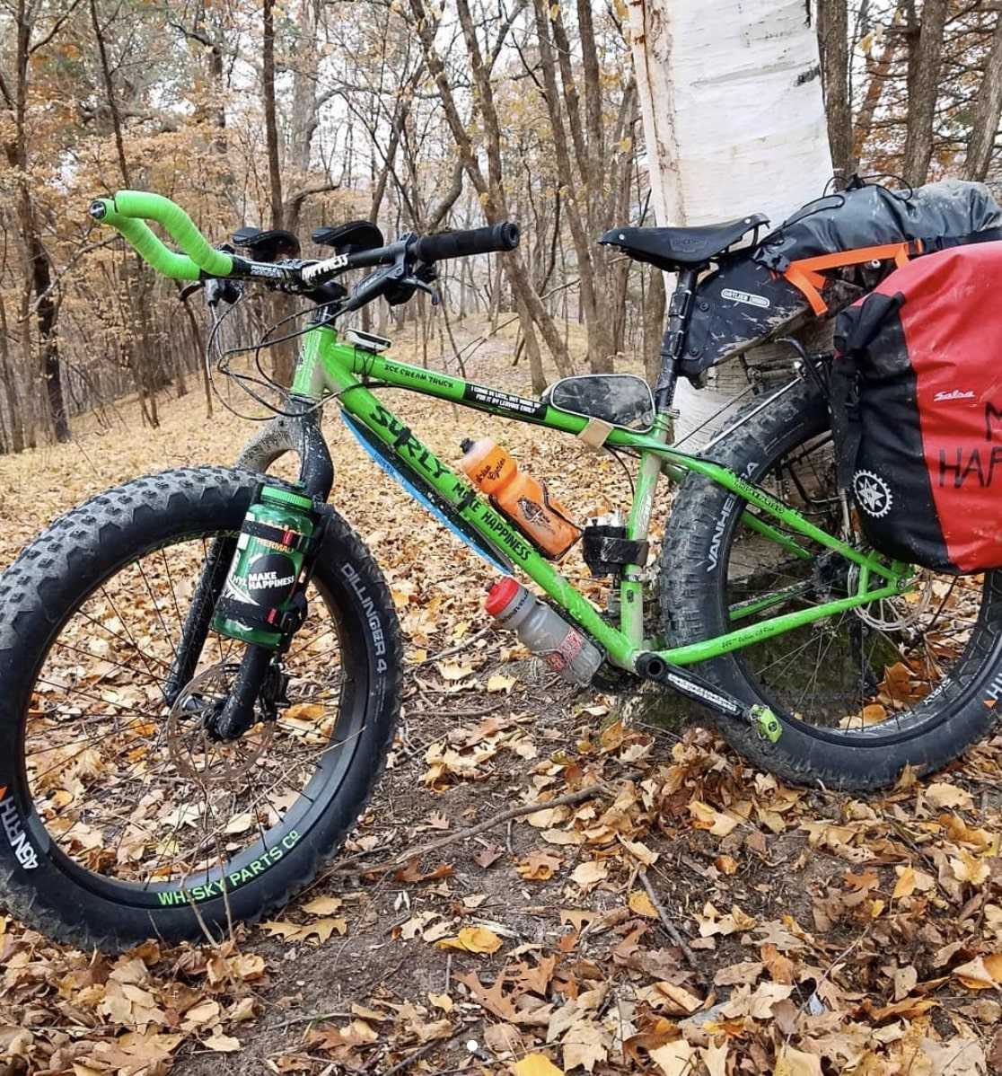 Left profile view of a green Surly Ice Cream Truck fat bike with rear saddle bags on leafy dirt trail in the woods