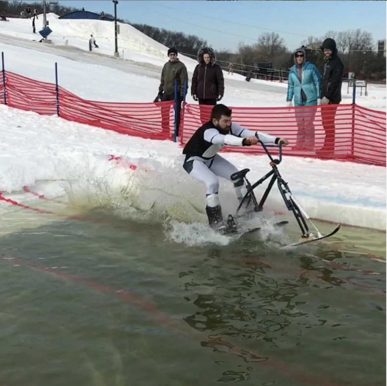 Person rides a bike with skis over a water hole in the snow at ski hill with spectators and a snow fence in background 