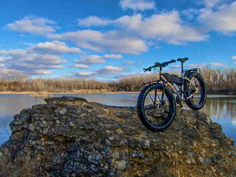 Left front view of a Surly Pugsley bike parked on a rock , with a lake and trees in the background