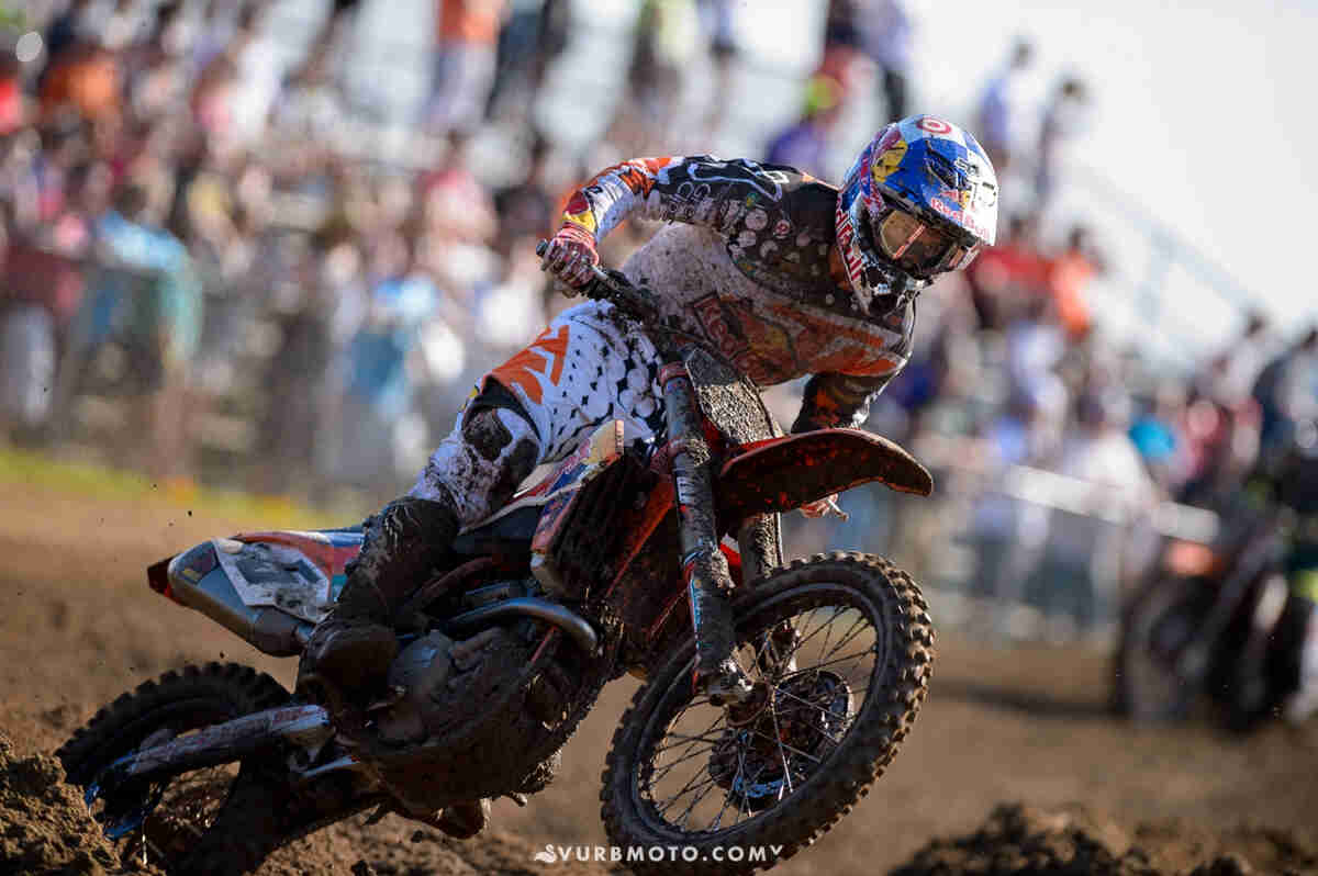 Right side view of a motocross racer, wearing a helmet and racing gear, rounding a turn on a dirt track