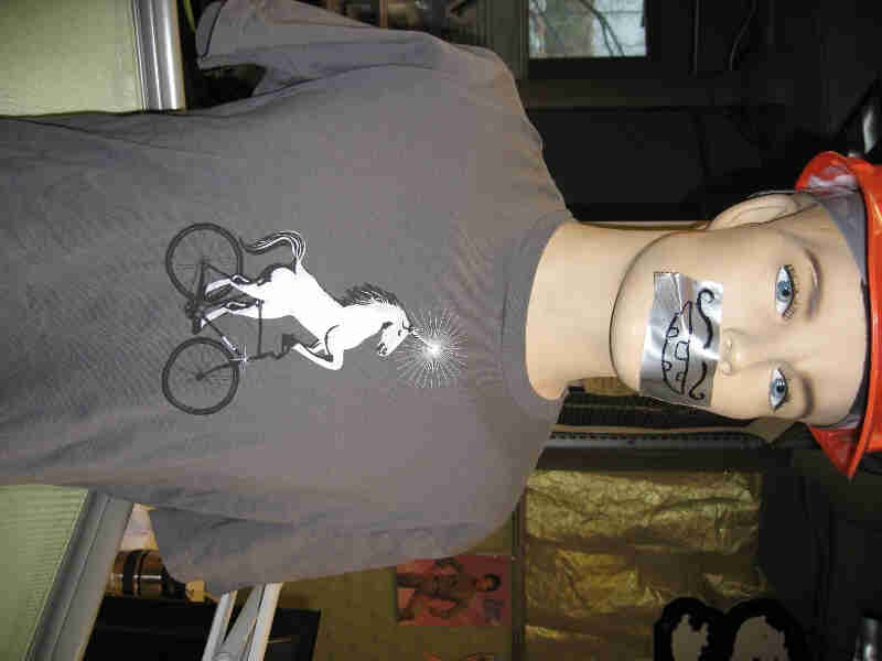 Waist up view of mannequin, wearing a gray Surly Bikes t-shirt with graphic of a unicorn riding a bike
