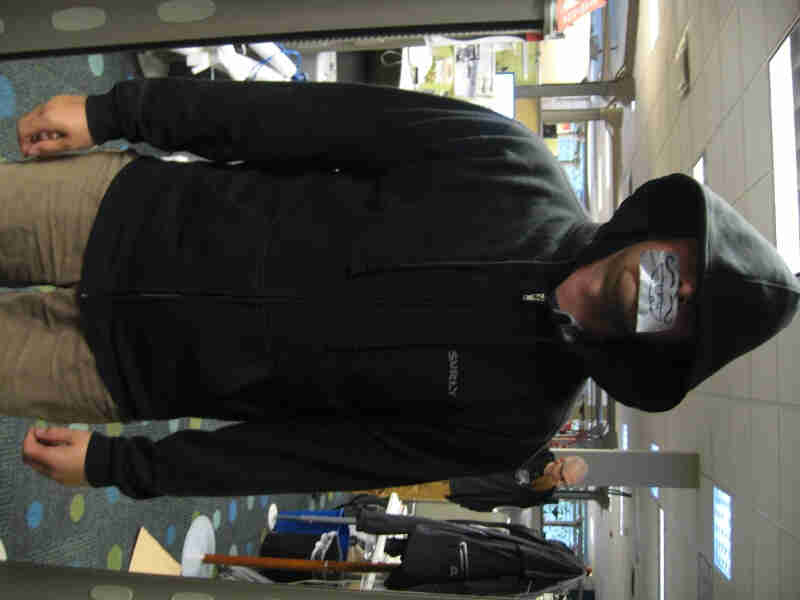 Front view of a person, wearing a black Surly Bikes Full Zip Hoodie, standing in an office with duct tape on their mouth