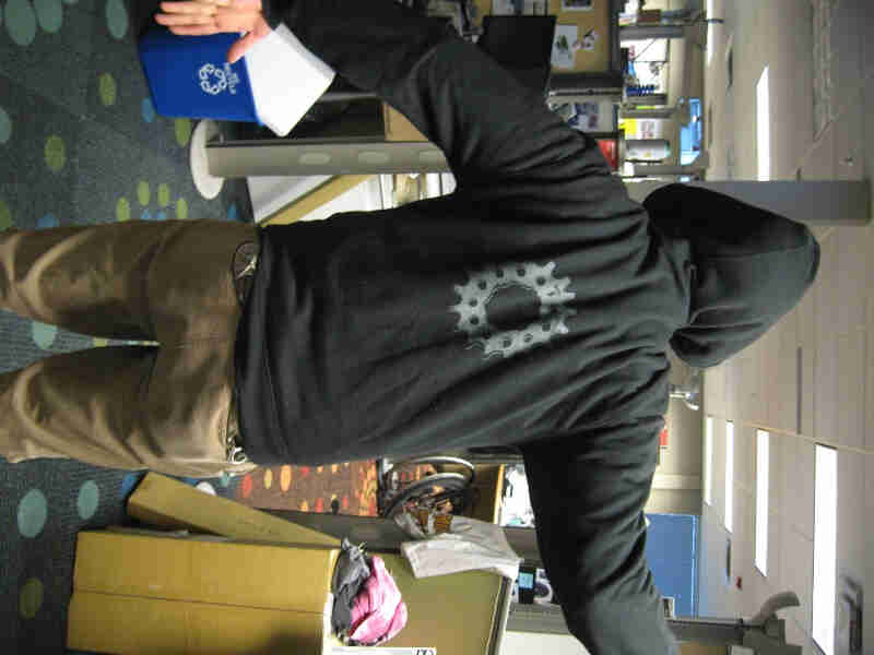 Rear view of a person, wearing a black Surly Bikes Full Zip Hoodie with a bike gear graphic, standing in an office