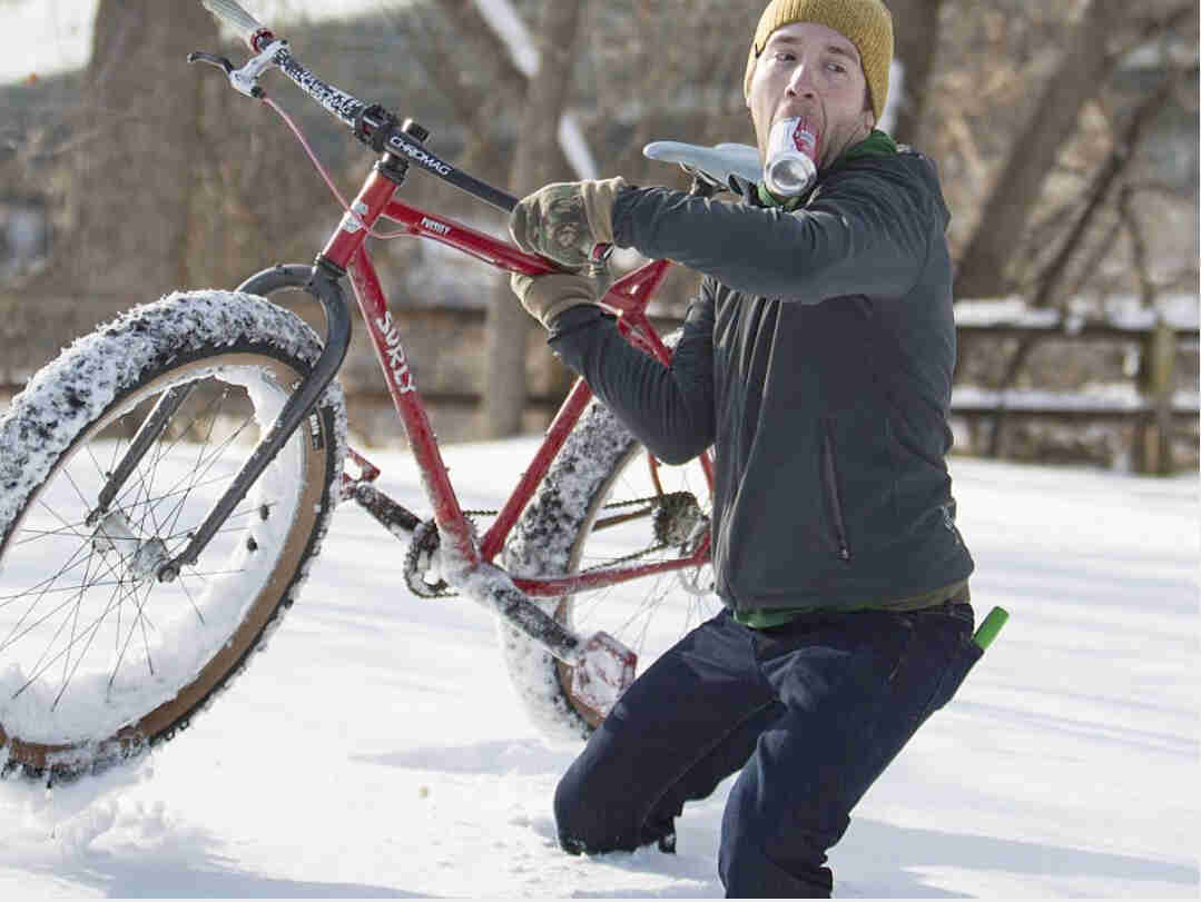 A cyclist, with a can in their mouth, carrying a red Surly Pugsley fat bike through knee deep snow