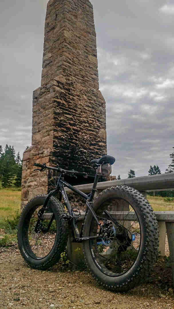 Rear left side view of a Surly Ice Cream Truck fat bike, black, parked on gravel in front of an outdoor stove with stack