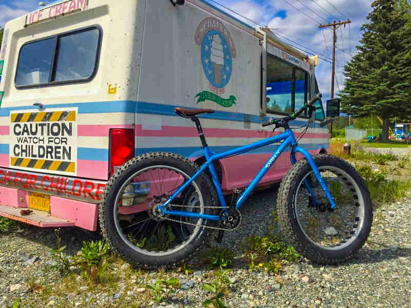 Right side view of a Surly Ice Cream Truck fat bike, blue, parked on gravel next to an Ice Cream Truck vehicle