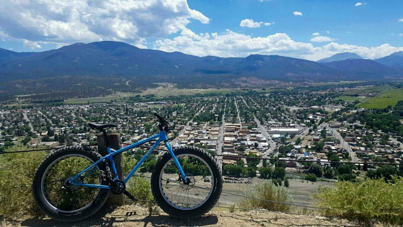 Right side view of a Surly Ice Cream Truck fat bike, blue, parked on hilltop, and a city with below with mountain behind