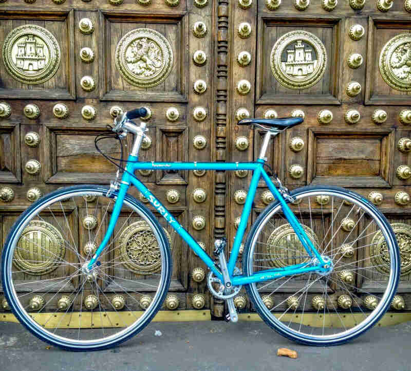 Left profile of a teal Surly bike, standing on pavement, in front of 2 large wood door with gold accents attached