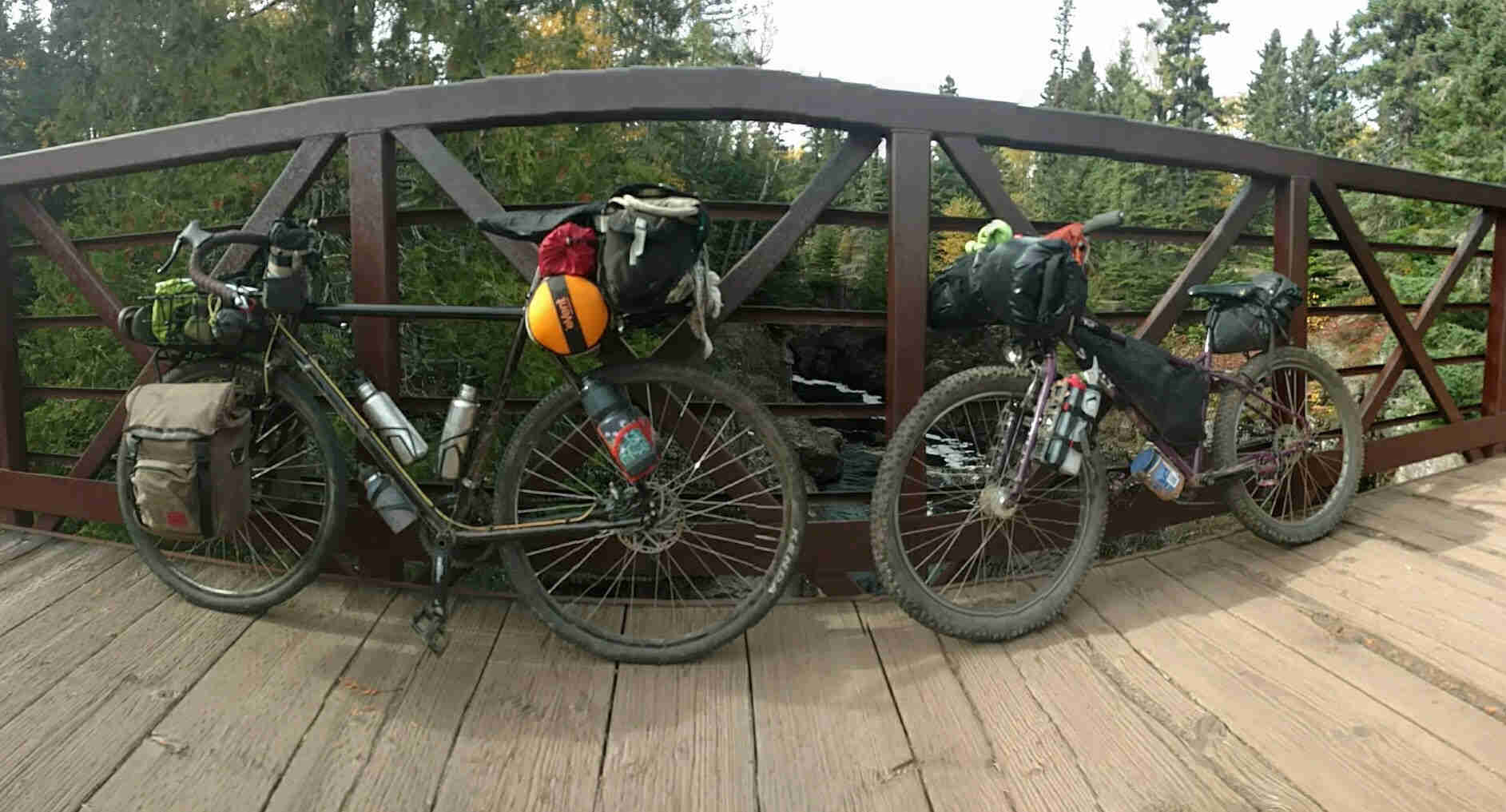 Left side view of 2 Surly bikes, loaded with gear, leaning on steel rail of a wood bridge over a river with trees surrounding it
