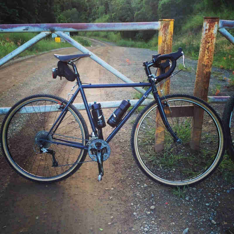 Right side view of a blue Surly Cross Check bike, parked on a gravel road in front of a closed gate