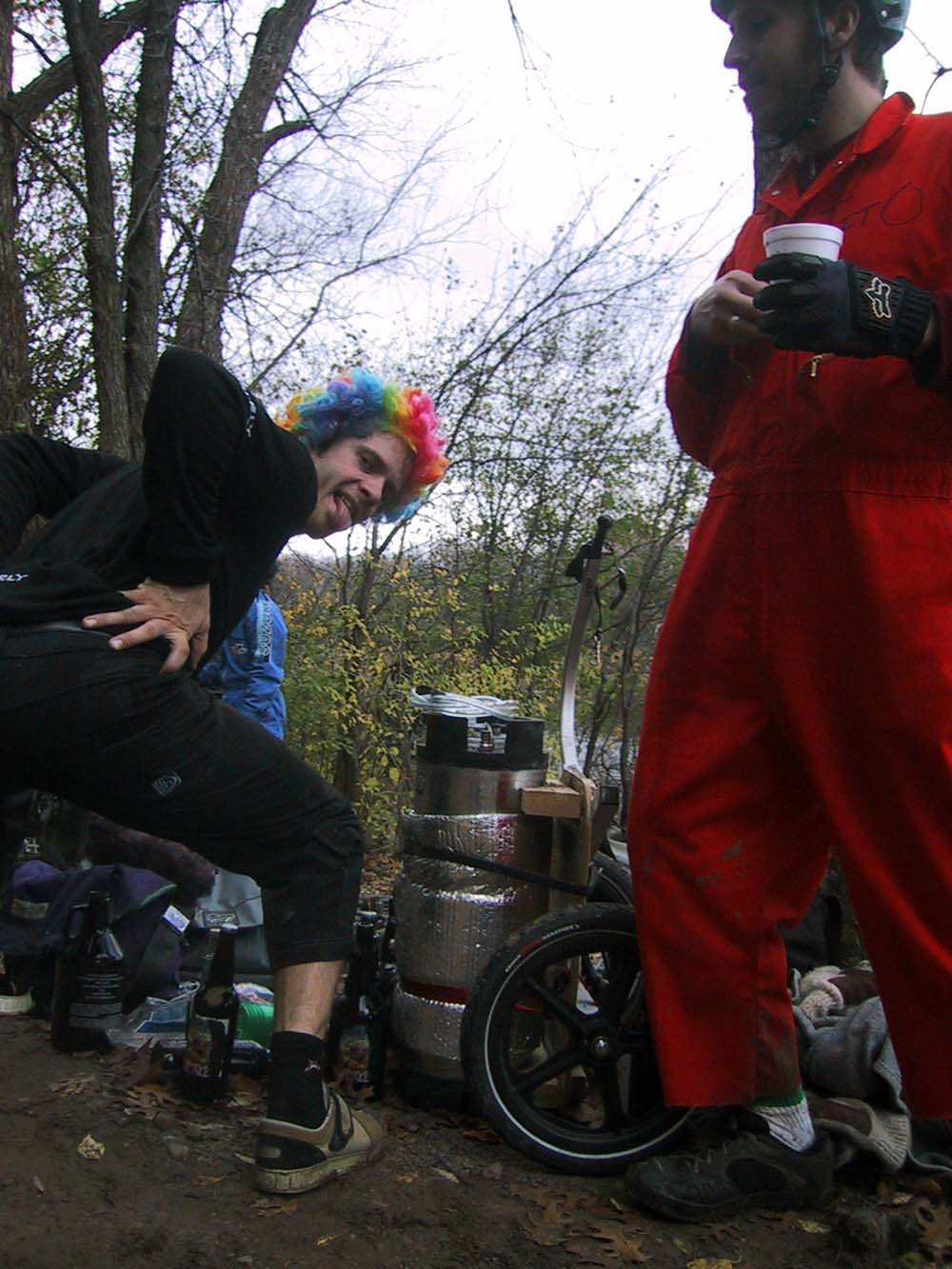 A person wear a rainbow wig and showing their rear end, next to a keg and a person in a red jump suit, in the woods