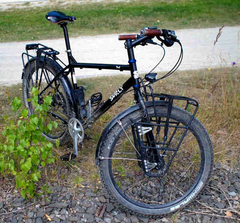 Front view of a black Surly Troll bike with it's front wheel turned to the left, standing on grass and rocks