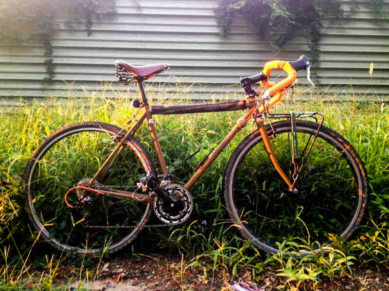 Right side view of an orange/purple Surly Cross Check bike, parked along tall weeds, with a steel wall in the background