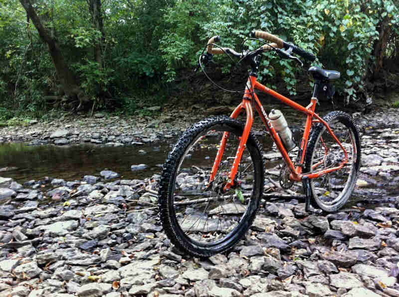 Front left side view of an orange Surly Troll bike, on the rocks of a stream bed, with the green woods in the background