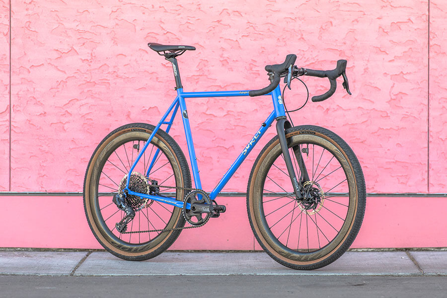 Right side view of a Surly Midnight Special Bike - Perry Winkle’s Sparkle - on a sidewalk alongside a pink stucco wall