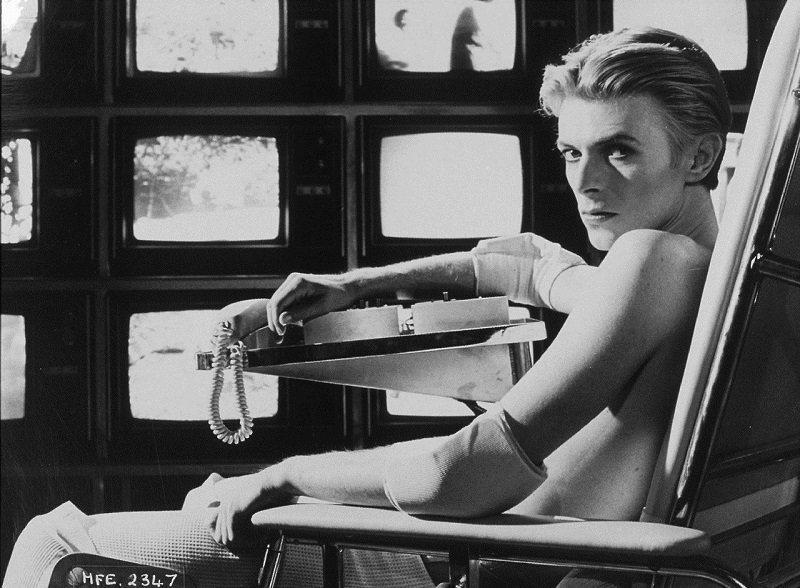 Black and White photograph of David Bowie sitting in a chair with a wall of TVs in the background