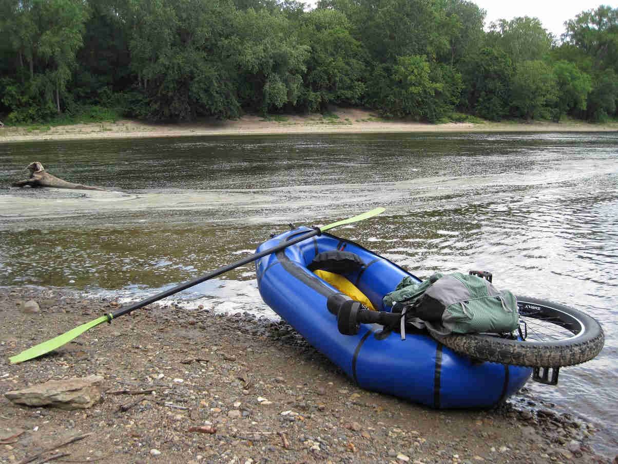 Rear view of a blue inflatable raft with an oar leaning on it, and a fat wheeled unicycle in the back, on a river bank