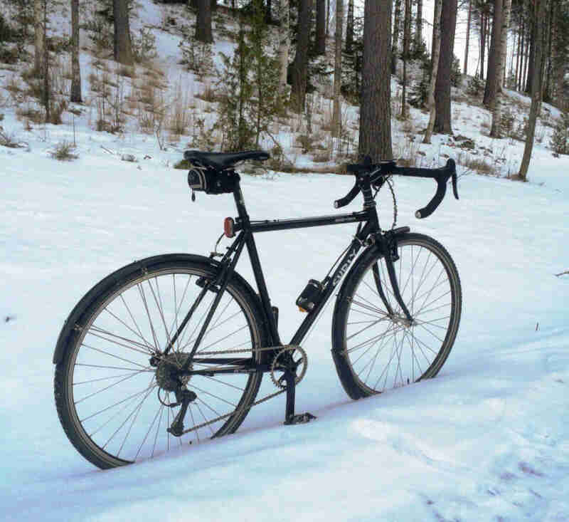 Right side view of a black Surly Cross Check bike, parked in a snowy trail, with a snowy forest hill in the background