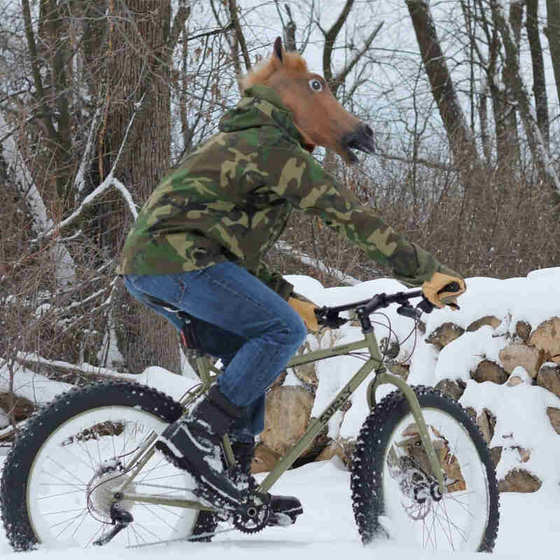 Ride side view of a cyclist, wearing a costume horse head, riding a tan Surly Pugsley fat bike on snowy ground