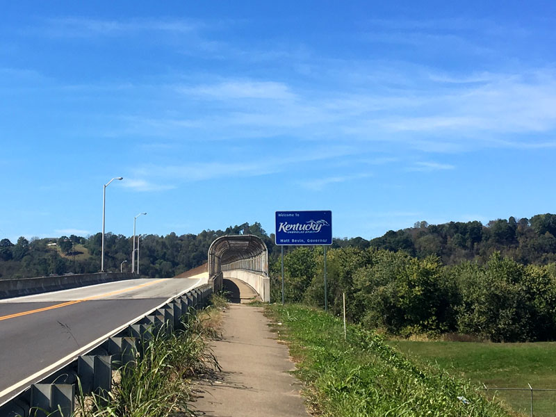 2 lane highway leading to a bridge with a bike trail and a Kentucky welcome sign on the right side with blue sky above